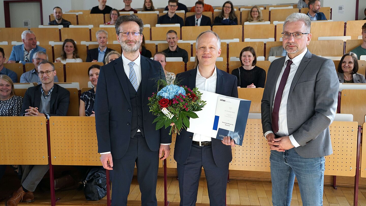 Gruppe von drei Herren mit Urkunde und Blumen vor Publikum in Hörsaal